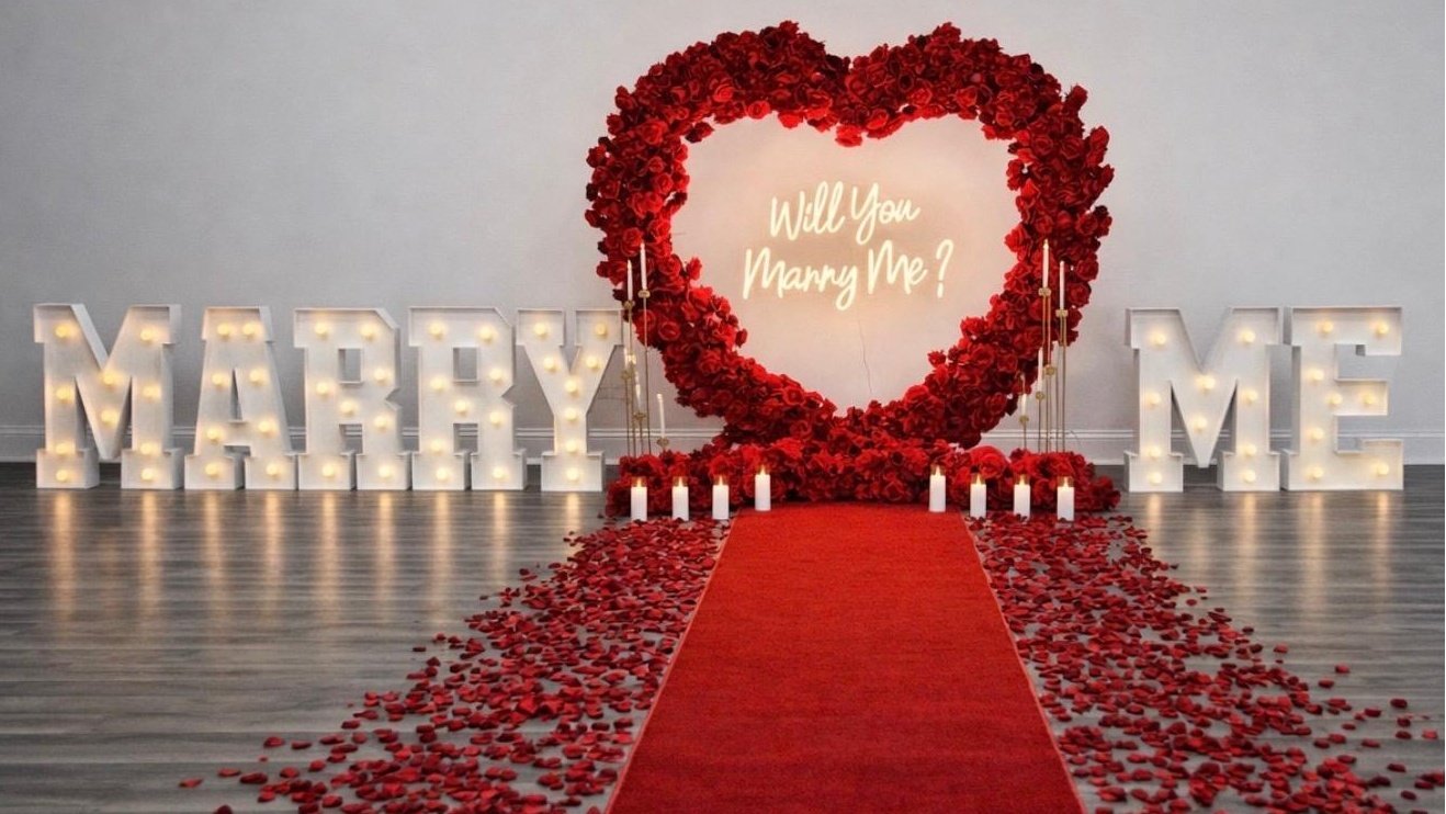 Red heart floral arch with Will You Marry Me neon sign, candles and rose petals